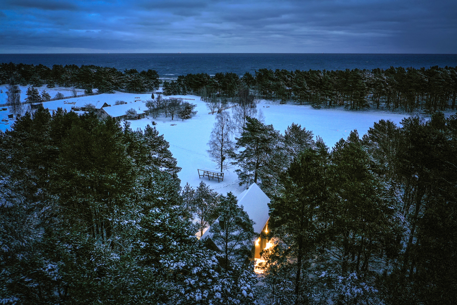 Brūna divstāvīga viesu māja ar gaismu logos no drona ziemā ar kokiem un jūru tālumā