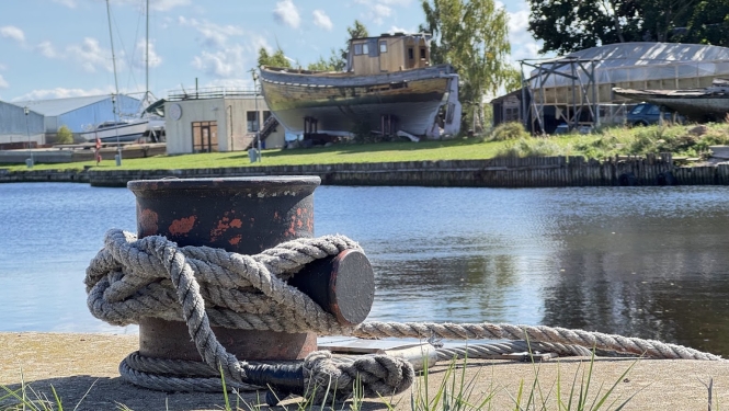 Bolderis kuģu piestātnē, fonā ūdens, pāri tam krastā izvilktas laivas