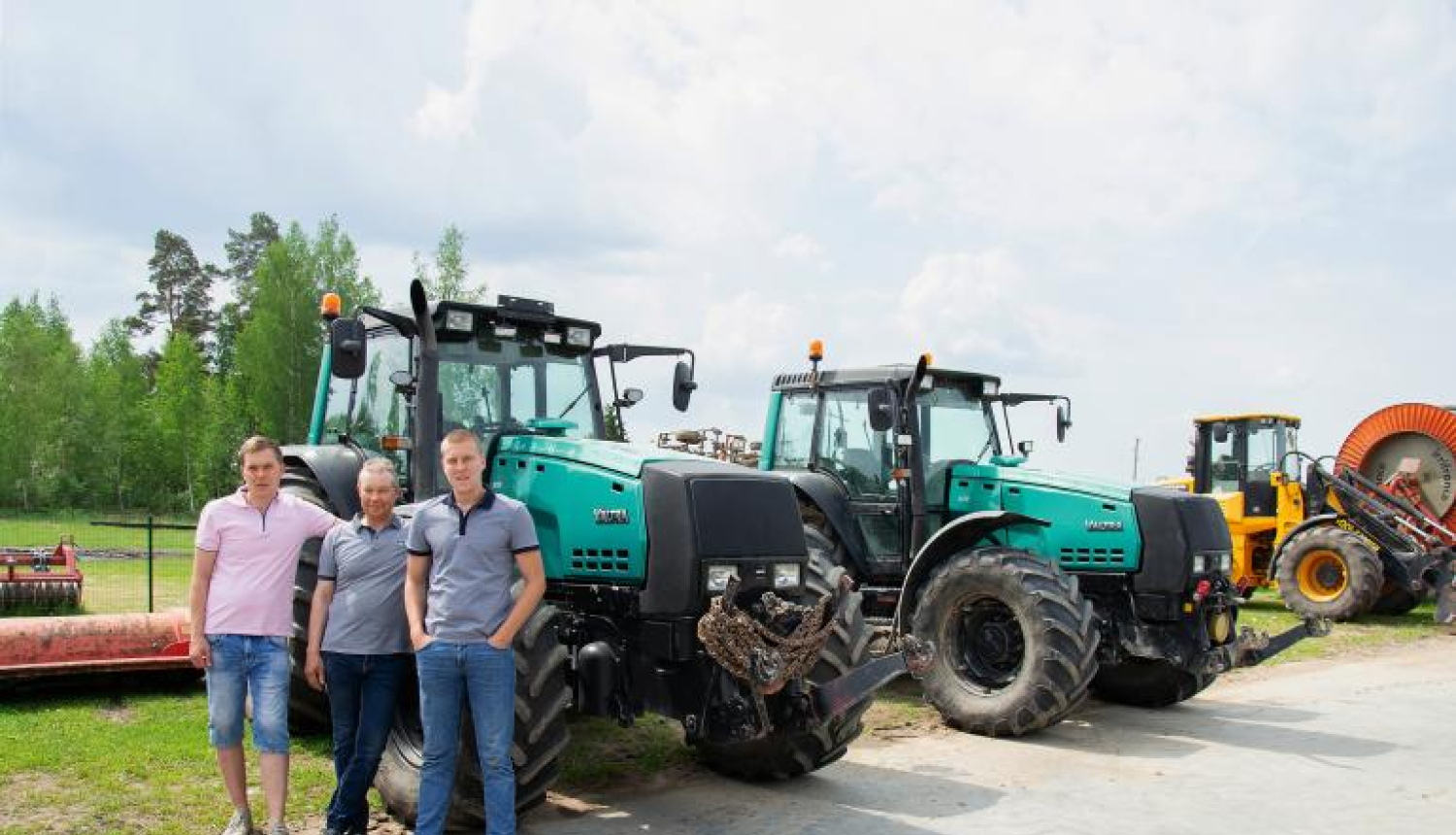 SIA Jāņlejas saimnieki stāv pie zaļa traktora, fonā vēl viens zaļš traktors un tālāk dzeltens traktors