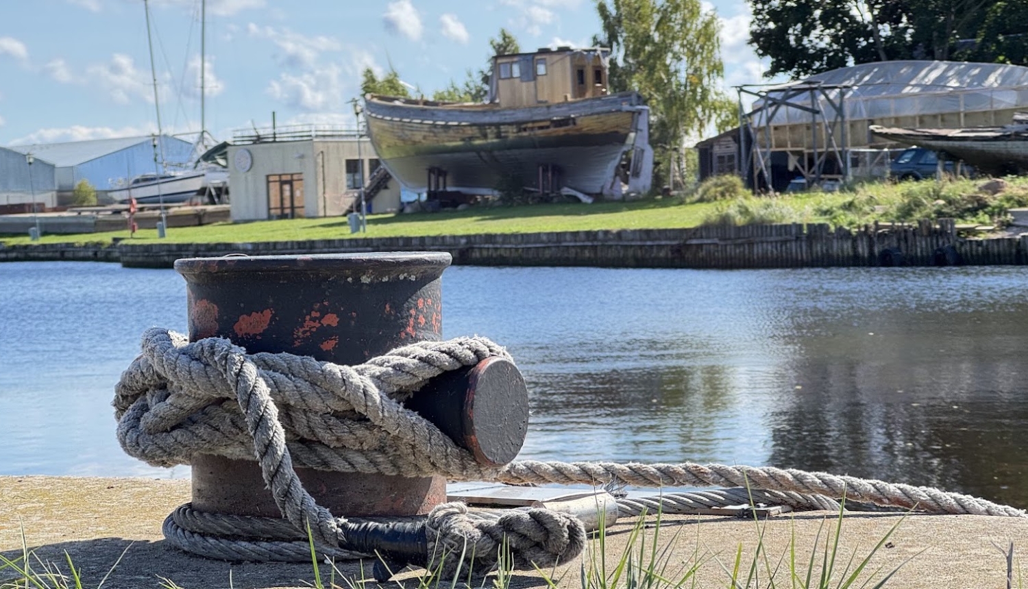 Bolderis kuģu piestātnē, fonā ūdens, pāri tam krastā izvilktas laivas