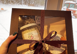 Rokās tiek turēts dāvanu komplekts ar saimniecības "Aronijas" produkciju. Fonā sniegots skats ārā pa logu.