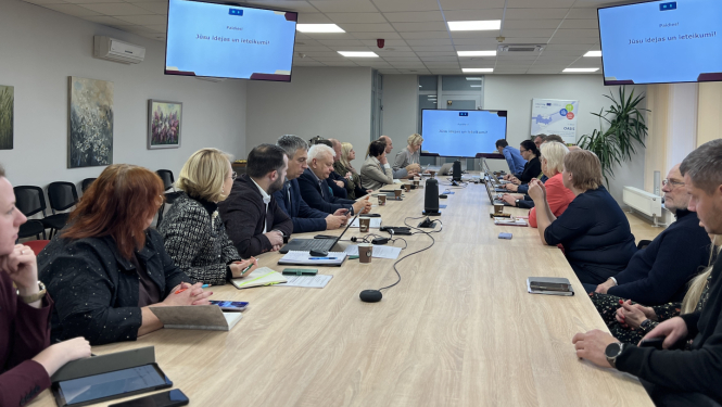 Cilvēki sēž pie gara galda un skatās uz ekrānu galda galā