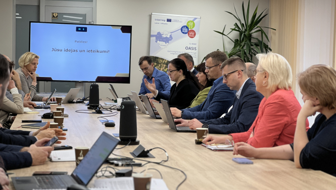 Pie gara galda sēž cilvēki, galda galā ekrāns, uz kuru visi skatās