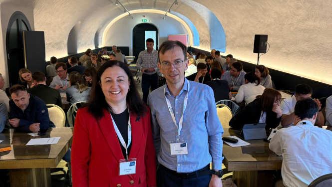 Divi cilvēki stāv priekšplānā, fonā konferences dalībnieki