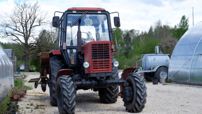 Lauksaimniecības Traktors ar piekabi stāv uz grants ceļa starp siltumnīcām un augiem podos.