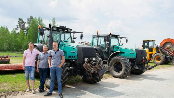 SIA Jāņlejas saimnieki stāv pie zaļa traktora, fonā vēl viens zaļš traktors un tālāk dzeltens traktors