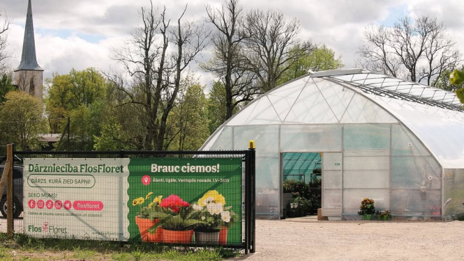 siltumnīcas ieeja, tās priekšpusē reklāmas plakāts ar puķēm, fonā koki un baznīcas tornis