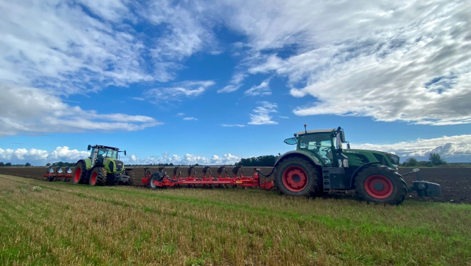 Divi zaļi traktori viens aiz otra ar zemi skaidrā saulainā dienā