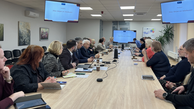 Cilvēki sēž pie gara galda un skatās uz ekrānu galda galā