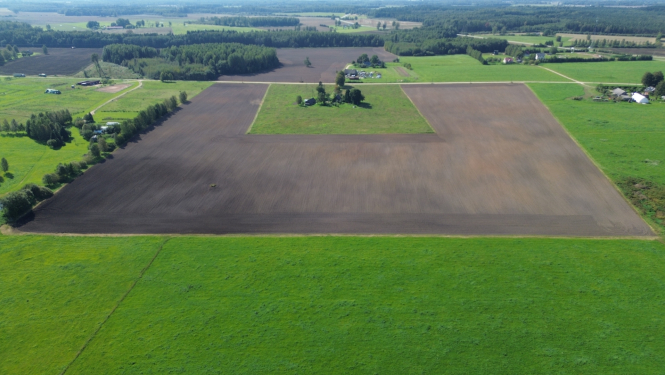 Zaļš lauks no putna lidojuma ar u veida apartu lauku un lauku māju pa vidu