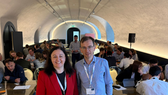 Divi cilvēki stāv priekšplānā, fonā konferences dalībnieki