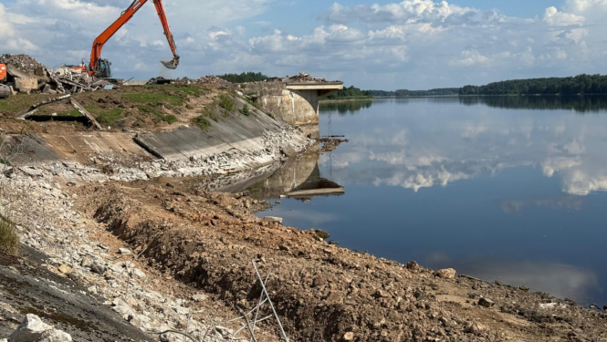 augsts upes krasts uz kura stāv sarkans ekskavators