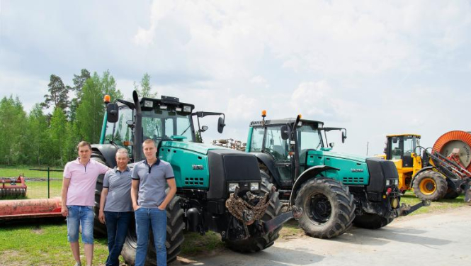 SIA Jāņlejas saimnieki stāv pie zaļa traktora, fonā vēl viens zaļš traktors un tālāk dzeltens traktors
