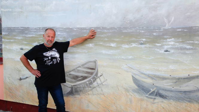 Zvejnieks stāv pie ējas, uz kuras sienas ir gleznojums ar jūru un laivām pludmalē