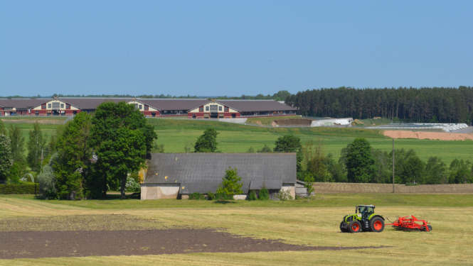 Skats uz lauku aimniecību - priekšpānā traktos apstrādā zemi, aiz tā veca saimniuecības ēka, pakalnā augšā jaunas fermas ēkas