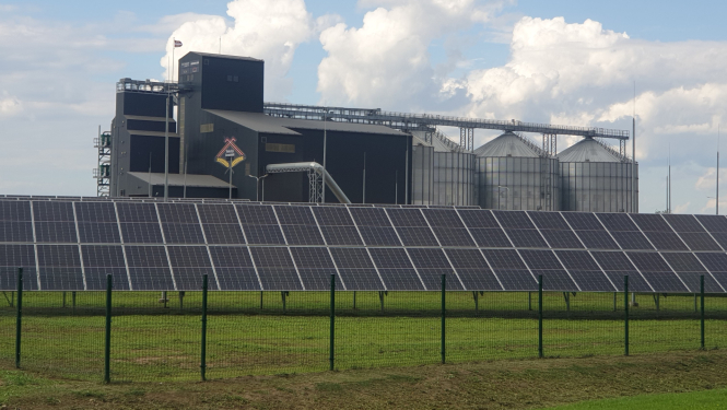 priekšplānā rinda ar saules paneļiem, aiz tiem graudu kalte ar torņiem