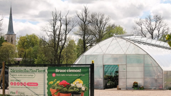siltumnīcas ieeja, tās priekšpusē reklāmas plakāts ar puķēm, fonā koki un baznīcas tornis