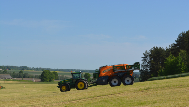 Zaļš traktors brauc uz leju pa nokalni, velk oranžu agregātu
