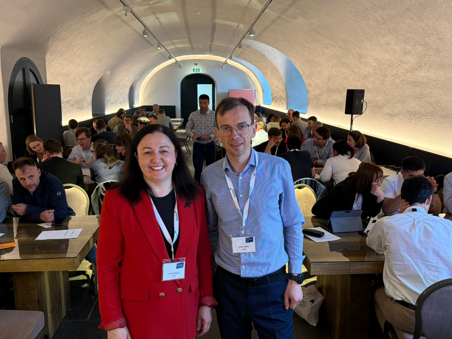 Divi cilvēki stāv priekšplānā, fonā konferences dalībnieki