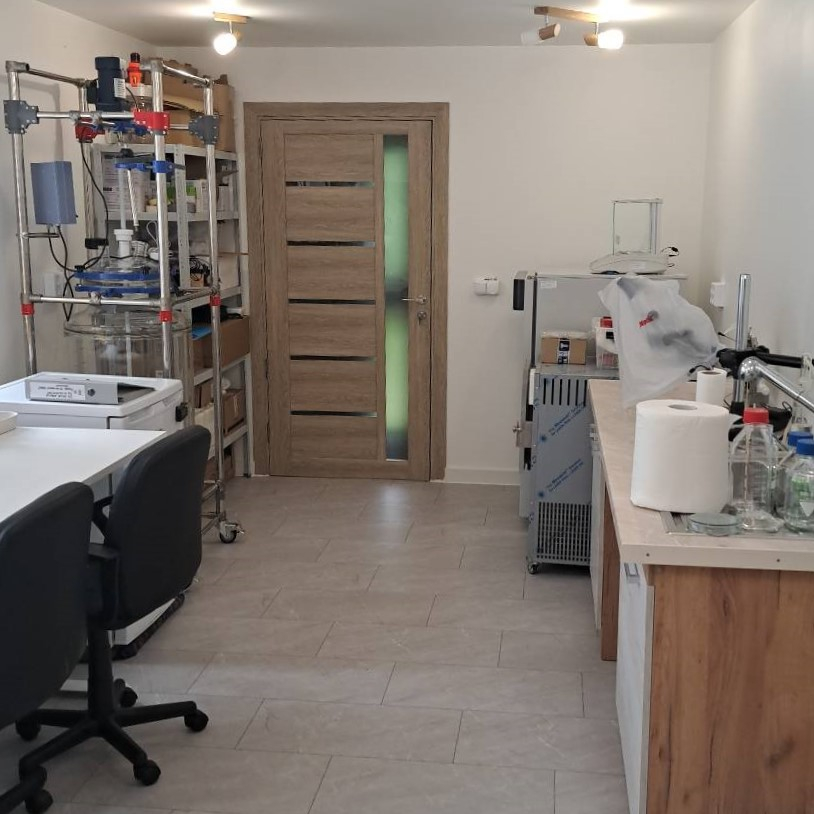 Labaratorijas skats, abās pusēs telpai galdi ar inventāru, telpas galā durvis