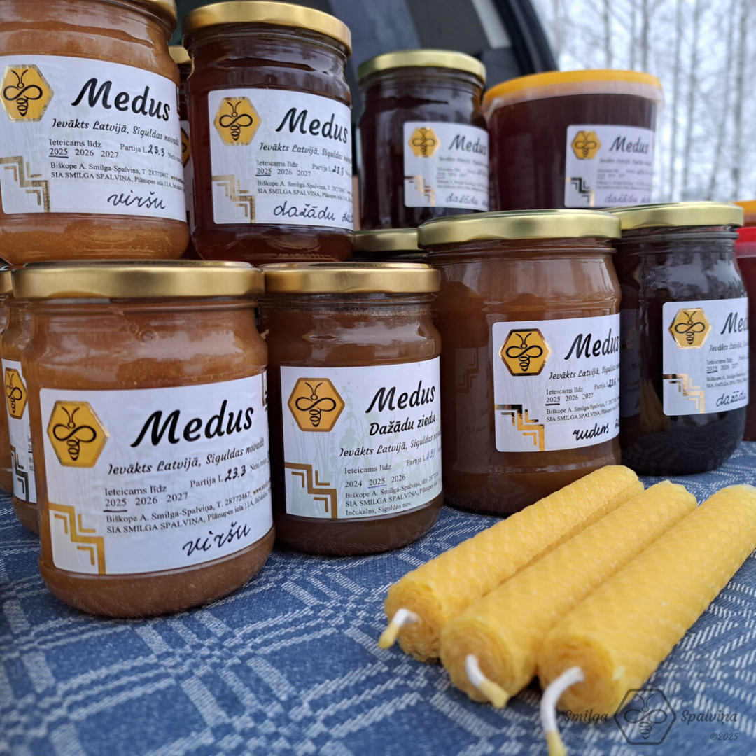 Medus burciņas izkārtotas divos stāvos, priekšpusē trīs vaska sveces