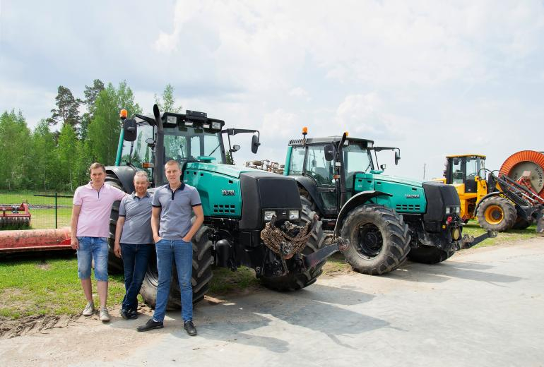 SIA Jāņlejas saimnieki stāv pie zaļa traktora, fonā vēl viens zaļš traktors un tālāk dzeltens traktors
