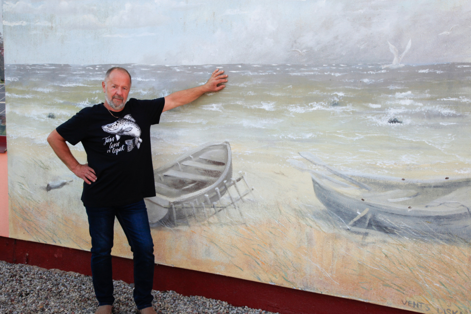 Zvejnieks stāv pie ējas, uz kuras sienas ir gleznojums ar jūru un laivām pludmalē