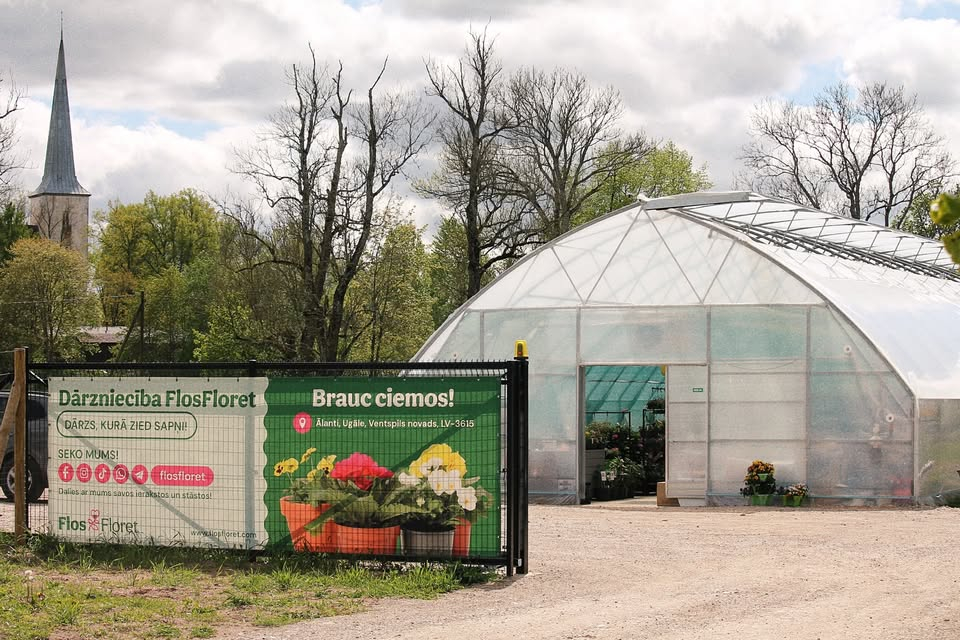 siltumnīcas ieeja, tās priekšpusē reklāmas plakāts ar puķēm, fonā koki un baznīcas tornis