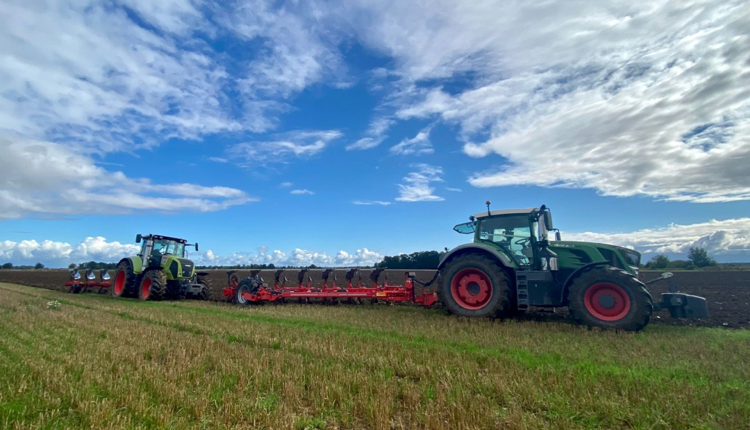 Divi zaļi traktori viens aiz otra ar zemi skaidrā saulainā dienā