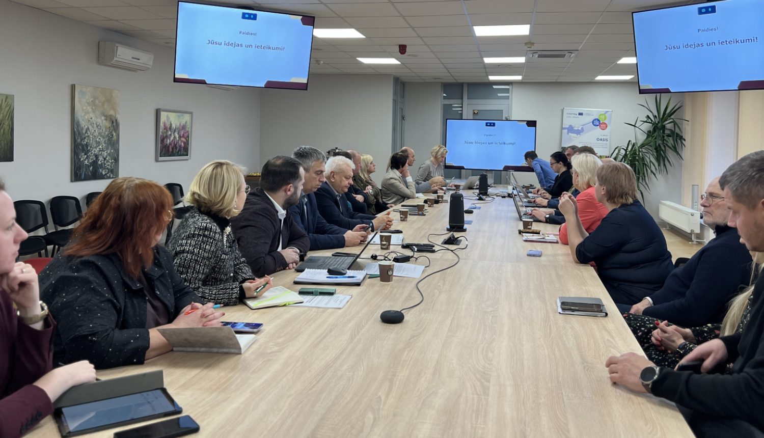 Cilvēki sēž pie gara galda un skatās uz ekrānu galda galā