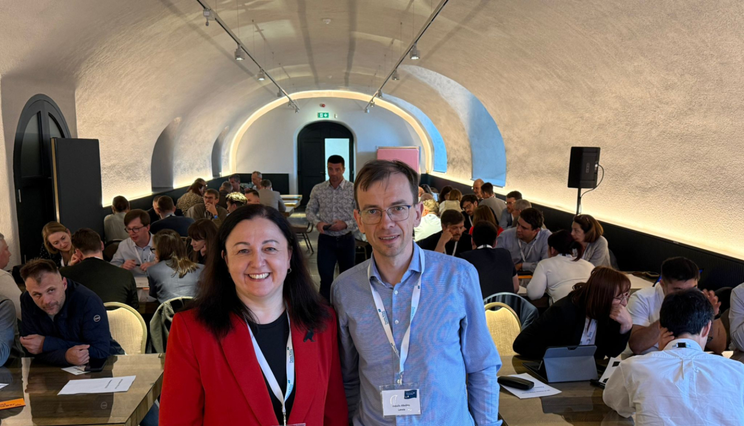 Divi cilvēki stāv priekšplānā, fonā konferences dalībnieki