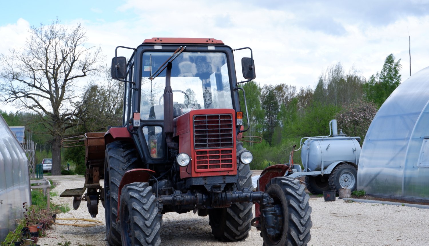 Lauksaimniecības Traktors ar piekabi stāv uz grants ceļa starp siltumnīcām un augiem podos.