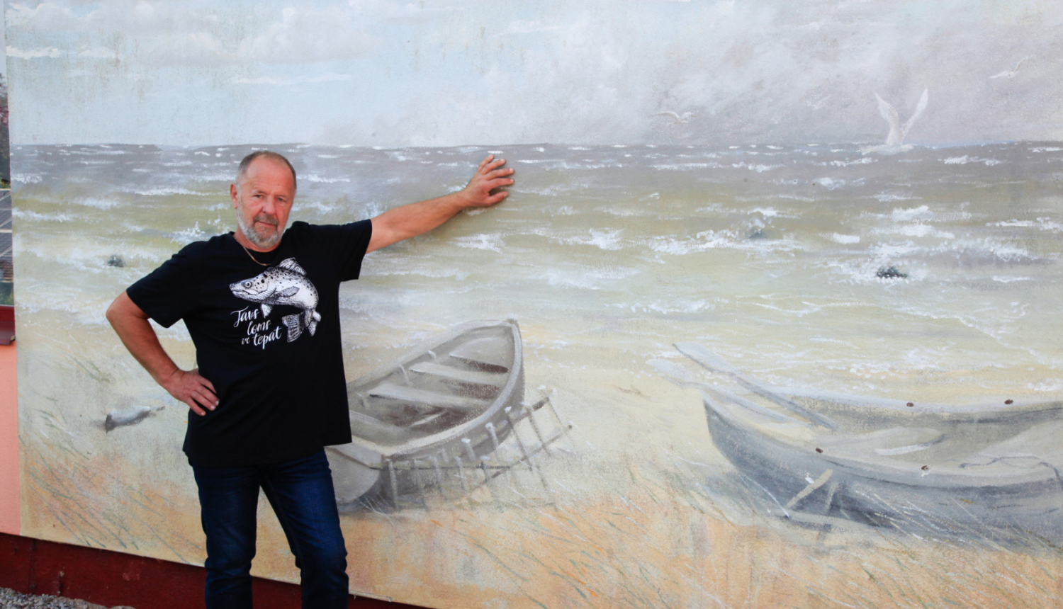 Zvejnieks stāv pie ējas, uz kuras sienas ir gleznojums ar jūru un laivām pludmalē