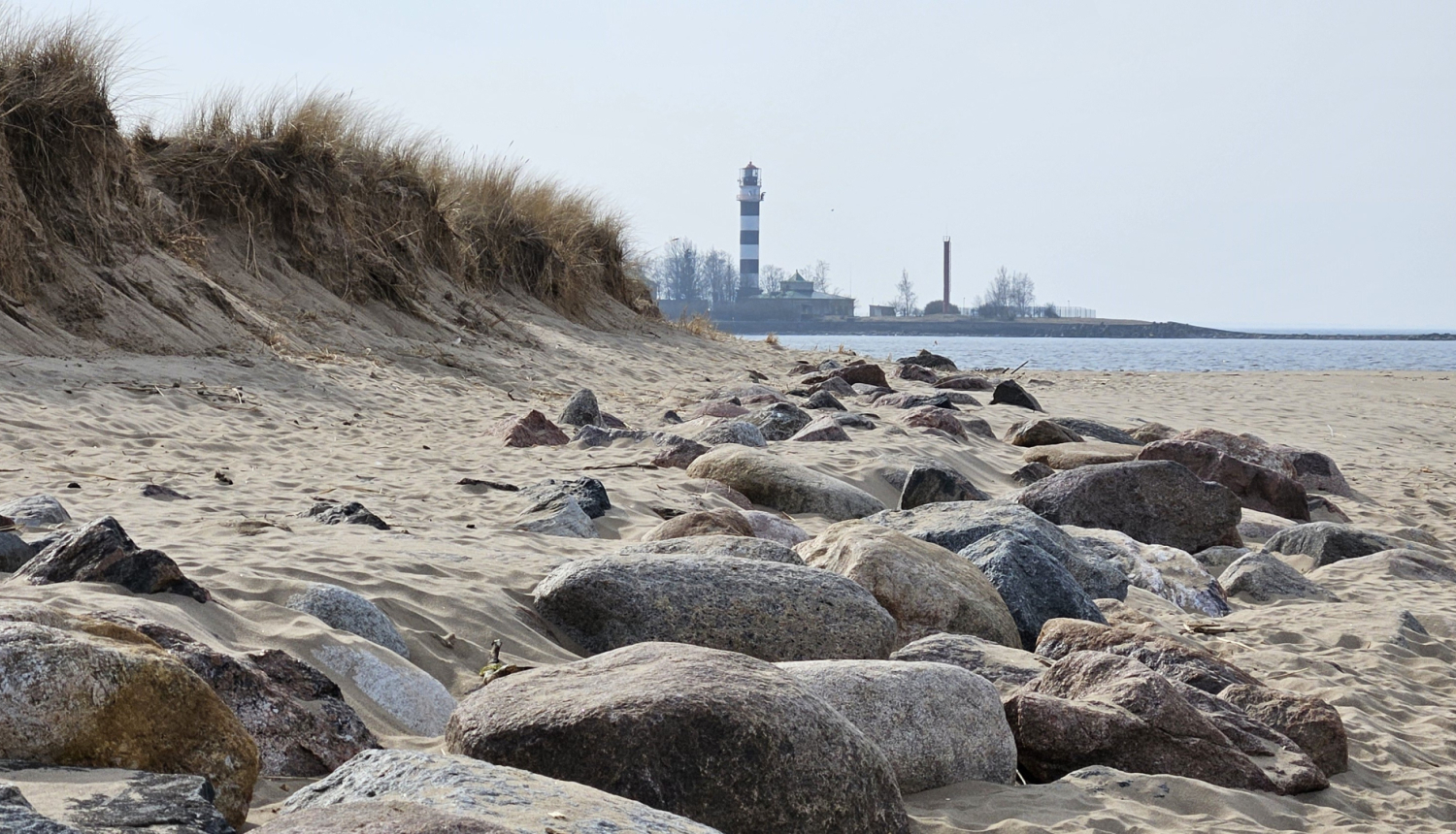 Jūras krasts, priekšplānā akmeņaina pludmale, tālumā bāka