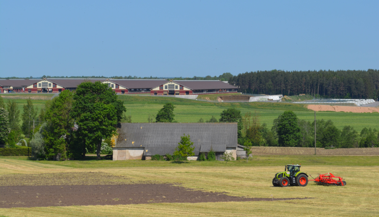 Skats uz lauku aimniecību - priekšpānā traktos apstrādā zemi, aiz tā veca saimniuecības ēka, pakalnā augšā jaunas fermas ēkas