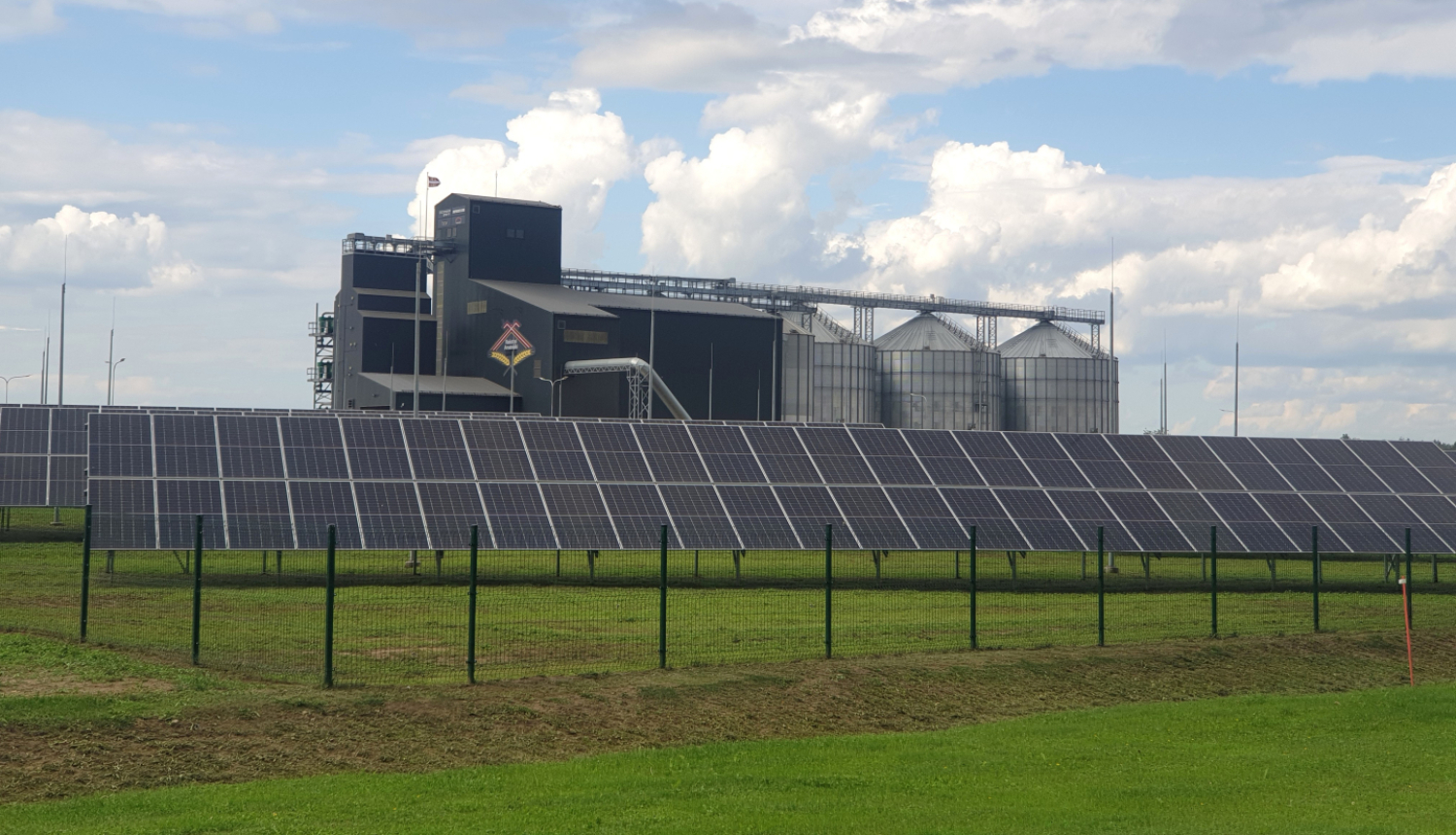 zaļā laukā saules paneļu rinda, aiz tiem fonā graudu uzglabāšanas torņi