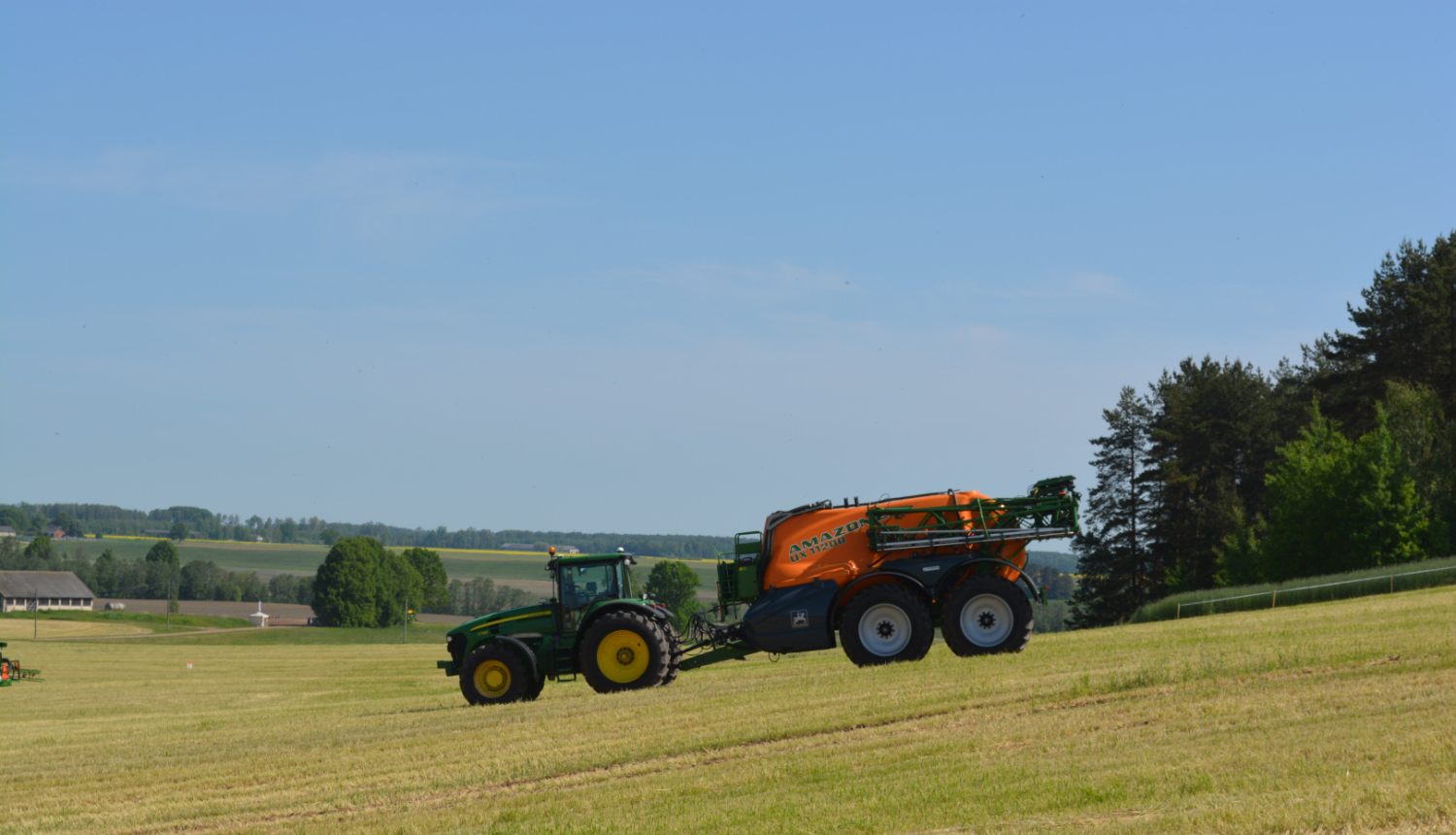 Zaļš traktors brauc uz leju pa nokalni, velk oranžu agregātu