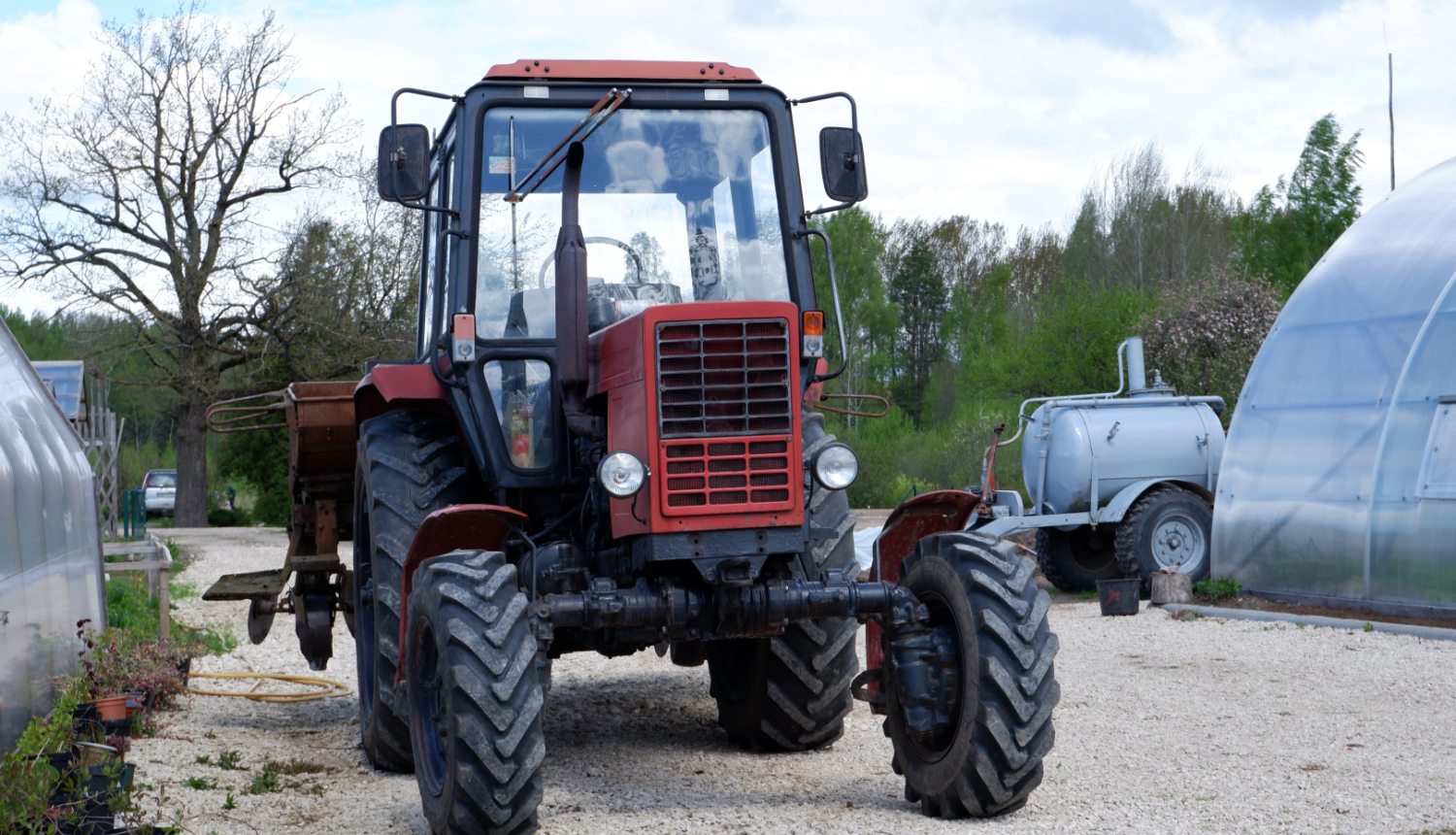 Lauksaimniecības Traktors ar piekabi stāv uz grants ceļa starp siltumnīcām un augiem podos.