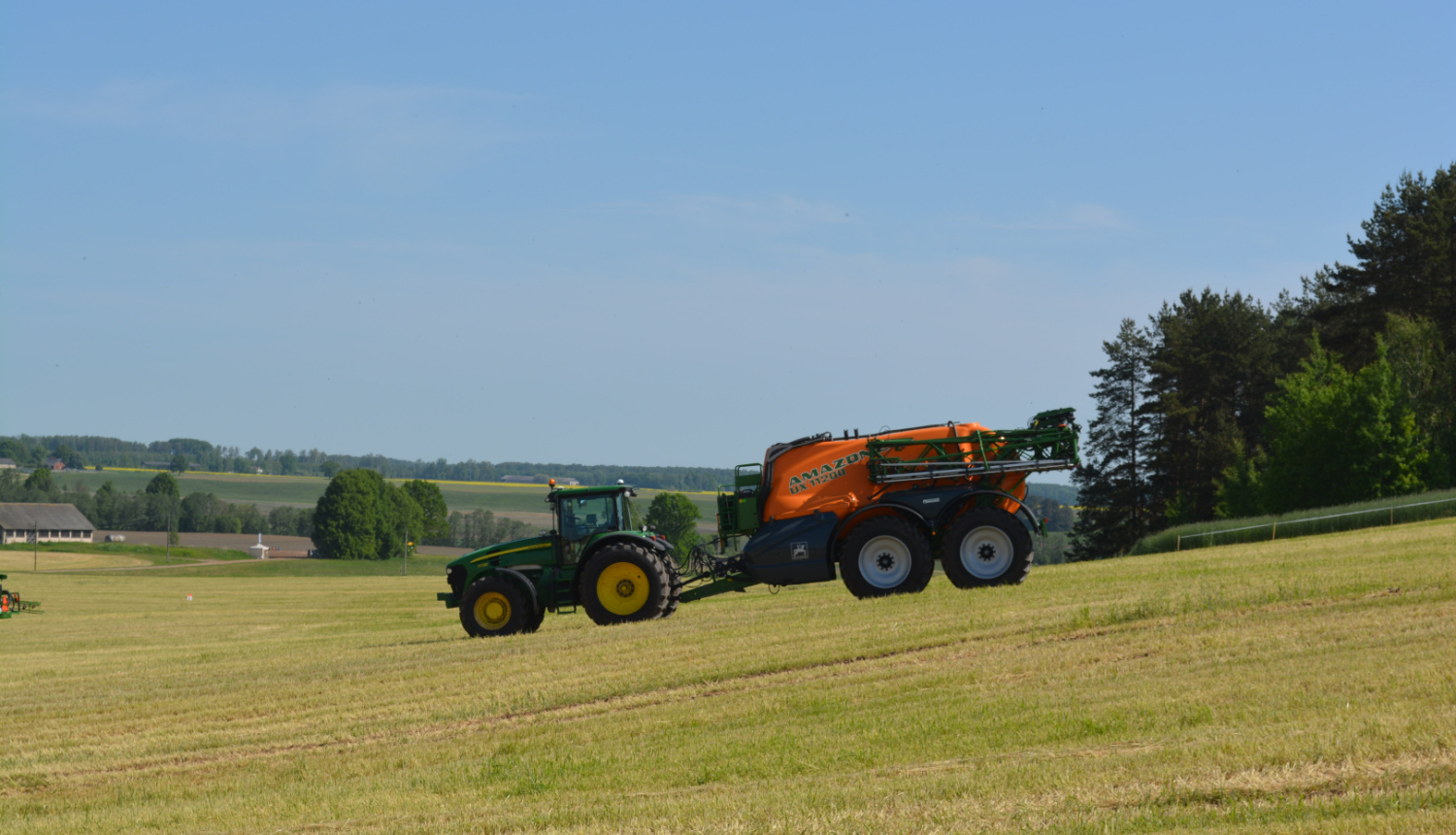 Zaļš traktors brauc uz leju pa nokalni, velk oranžu agregātu