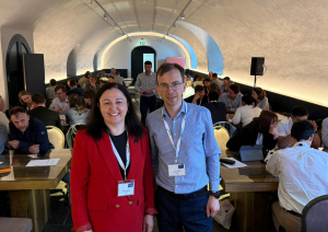 Divi cilvēki stāv priekšplānā, fonā konferences dalībnieki